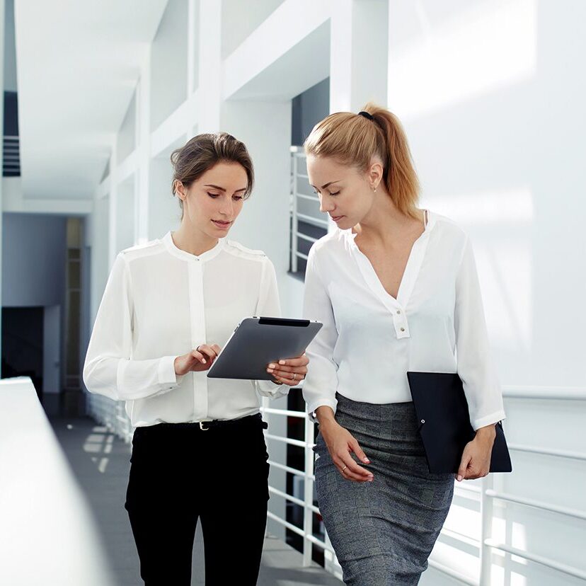 Two female managing directors discussing ideas of project on digital tablet while walking down in office hall, confident women entrepreneurs working on touch pad while going to the conference room